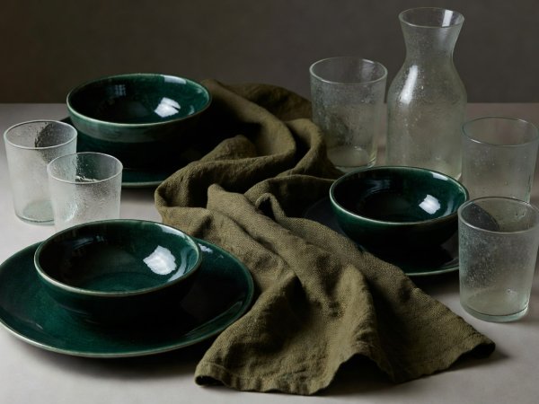 Composition of dark emerald glazed ceramics, coarse olive linen, and textured glassware catching soft side-light
